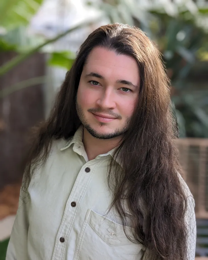 Jean-Luc Alder, smiling, with long dark hair and a pale green shirt, standing in a leafy garden.